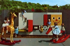 Children play in a vintage playground featuring a large colorful wall with puzzle elements and smiling face design. Kids are on a giraffe and horse rocker in the foreground, while others interact with the walls features. Trees form the background.