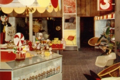 Vintage toy store interior featuring a red and yellow color scheme. A glass display case holds various toys. Signs with TOYS and a duck motif hang above. A red horse toy is in the foreground, and a large arrow points towards the back.