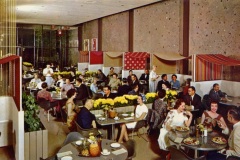 A mid-20th-century restaurant with patrons seated at tables, eating and socializing. The decor includes yellow flowers, colorful striped canopies, and a stone-textured wall. The ambiance is lively and bustling.