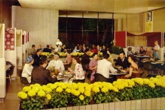 A vintage restaurant scene with numerous well-dressed patrons seated at tables eating and conversing. Yellow flowers in the foreground, partitioned booths, and large windows create a stylish, lively ambiance.