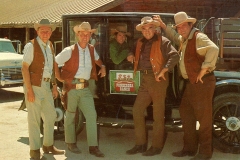 Five men in cowboy attire stand beside a vintage car labeled Ponderosa Ranch. They wear hats, vests, and holsters, and are posed casually, evoking a classic Western scene. A wooden building and an old car are in the background.