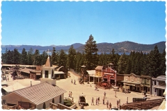 A vintage postcard depicts the Ponderosa Ranch, a Western-style town with a church, colorful storefronts, and people walking along the streets. Tall pine trees surround the area, with a scenic lake and mountains visible in the background under a clear blue sky.