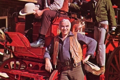 Three men in vintage Western attire posing with a red horse-drawn fire engine. One sits on top wearing a fire helmet, another leans on the wheel, and the third stands behind, all smiling in front of a wooden building.