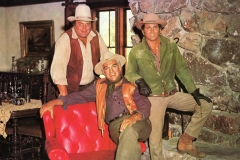 Three men, the Cartwrights, Hoss,Ben and Little Joe,  in cowboy attire pose in a rustic room. One sits on a red chair, while the others stand nearby. They wear traditional cowboy hats and vests, and a stone wall is visible in the background.