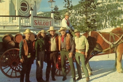A group of six people dressed in Western attire stand in front of the Silver Dollar Saloon. Behind them is a horse-drawn wagon with barrels. Snow covers the ground with pine trees and a building in the background.