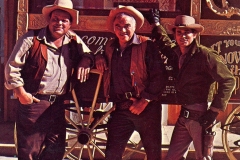 Three men in cowboy attire stand in front of a vintage wagon with the words Ponderosa Ranch Mountain Dew painted on it. They are wearing hats, vests, and boots, smiling at the camera in a Western-style setting.