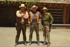At the Ponderosa Ranch, Three men dressed in cowboy attire stand in front of a rustic wooden building. They wear wide-brimmed hats, vests, and belts with holsters. The background shows a wooden structure with a shingled roof.