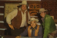 Three men in cowboy attire stand and sit indoors. The man in the center is seated at a table, while the other two stand beside him. They are wearing hats and vests, and behind them, there are guns mounted on the wall and a map.