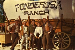 Five people stand in front of a large covered wagon with Ponderosa Ranch written on it. They are wearing cowboy attire, including hats and boots, giving a western theme. The background includes a wooden structure under a clear sky.