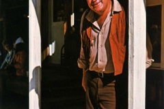 A man wearing a cowboy hat, vest, and pants casually leans against a wooden post outside the Silver Dollar Saloon. He is smiling, with one foot resting on the post, capturing a relaxed Old West atmosphere.