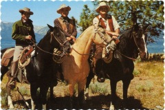 Three cowboys on horses pose outdoors. They wear hats and western attire. The backdrop features trees and a glimpse of a lake or mountains, suggesting a Western setting. The image has a vintage, postcard-like appearance with a scalloped border.