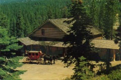 A rustic log cabin surrounded by dense green forest. In front, a red stagecoach with two horses waits on a dirt path. The scene captures a serene, vintage atmosphere in a natural setting.