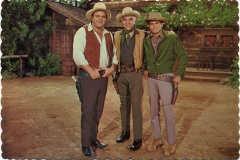 Three men in cowboy attire stand smiling in a rustic outdoor setting with wooden buildings and trees. They wear hats and have holstered guns, creating a classic Western scene.