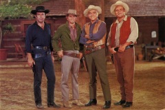 Four men dressed in cowboy attire stand together outdoors. They wear hats, belts with holsters, and boots. A wooden building is in the background, and they are on a dirt surface. Their expressions are friendly and relaxed.