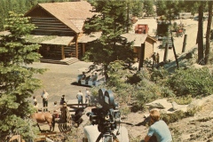 A film set with a log cabin surrounded by trees. Crew members and a camera are visible in the foreground, while several people and a horse stand near the cabin. A dirt path and equipment are seen in the background.