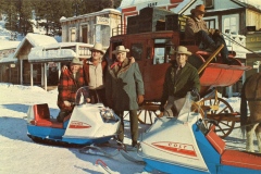 Four people in cowboy hats stand in the snow with two vintage snowmobiles in front of a stagecoach. Theyre in a western-style town with wooden buildings. One person is seated on the stagecoach. Snow covers the ground.