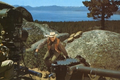 A cowboy in a hat and gloves acts in front of a film camera on a rocky set with large boulders. A pine forest and a blue lake with mountains are in the background under a clear sky.