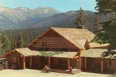 A rustic log cabin with a shingled roof surrounded by pine trees and mountains in the background. The scene is set on a sunny day, and theres a vintage car parked to the left of the cabin.