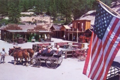 A Western-style village scene with a trading post, wooden buildings, and people in period clothing. Horse-drawn carriages are on a dirt road. A large American flag waves in the foreground under a clear blue sky.