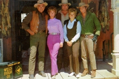 A group of five people in cowboy attire stand on a wooden porch. Men wear hats and Western clothing, and two women are dressed in colorful 1960s outfits. A Wells Fargo box and cowboy accessories are in the background.