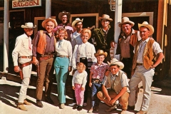 The Ponderosa Ranch hands. A group of people dressed in Western attire, including cowboy hats and boots, standing in front of a saloon. There are men, women, and children smiling at the camera under a sunny sky.
