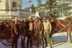 A group of five people in cowboy attire poses in front of a horse-drawn wagon with barrels. A saloon sign is visible in the background. Snow and pine trees surround the scene, suggesting a western or alpine setting.