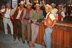 Seven men in cowboy attire, including hats and vests, stand and lean against a bar counter in a saloon setting. Drinks are visible on the counter, and all are smiling or laughing. The background features wood paneling and dim lighting.