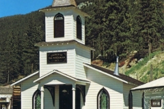 A small white wooden church with a steeple and a cross on top, surrounded by a white picket fence. Green trees and a clear blue sky form the background. The sign reads Church of the Holy Cross.