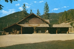 A rustic log cabin with a pitched roof stands against a backdrop of pine-covered mountains. The cabin has a wide porch, wooden barrels, and a gravel area in front, under a bright blue sky with scattered clouds.