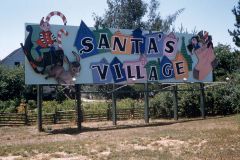 A colorful billboard in Skyforest, California, reads Santas Village with cartoon images of a cheery elf, a donkey, and a woman in a bonnet, set against houses and trees in a grassy outdoor area—celebrating its charm since in1958.