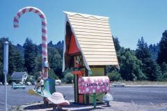 A whimsical roadside structure in Skyforest, California, featuring a large candy cane and an oversized mailbox labeled “Santa Claus,” with children climbing playful decorations amid trees and cars, reminiscent of holiday magic in1958.