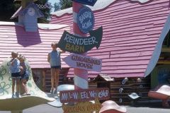 A whimsical signpost painted like a candy cane stands in front of playful buildings at Santa’s Village in Skyforest. The signs point to places like the North Pole and Mrs. Claus’ Kitchen, while children stand nearby on oversized, colorful toadstools.