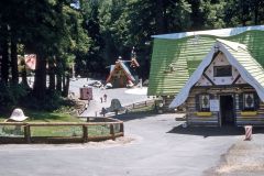 A whimsical park in Skyforest, California, with colorful, fairy-tale style houses, surrounded by tall trees. Children and adults stroll along paved paths and play near the houses on a sunny day in Santas Village.