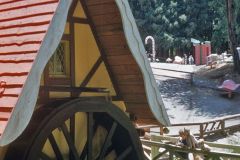 A large wooden watermill stands by a small stream with a red roof and yellow walls; trees and fairy tale-themed park structures mark this enchanting corner of Santas Village in Skyforest, California.