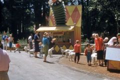 Children and adults gather near a whimsical booth topped with a large jester head at Santas Village in California. Some children ride toy cars, goats wander nearby, and trees and greenery surround the lively 1958 scene.
