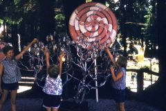Three children reach up to hang or retrieve keys from a silver tree beneath a large red-and-white spiral lollipop decoration at Santas Village in Skyforest, California, surrounded by trees in an outdoor, park-like setting.