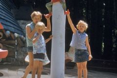 Three children stand around a tall white pole topped with a character in a North Pole shirt at Santas Village in Skyforest, California, surrounded by trees and whimsical decorations in this enchanting theme park setting.