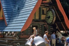 Children and adults stand in front of a whimsical house with a slanted blue roof and a large clock. A child sits on a mushroom sculpture near a stroller. This playful, outdoor scene could be from Skyforest, California in 1958.