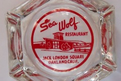 This hexagonal, clear glass ashtray showcases a striking red and white design. It proudly displays the name Sea Wolf Restaurant, accompanied by an illustration of a building and the iconic Jack London Square, Oakland, Calif., inion.