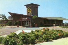 Vintage image of the Sea Wolf Restaurant at Jack London Square, showcasing its distinctive mid-century modern design. The wooden building features a tall sign and large windows, surrounded by a landscaped garden with vibrant flowers. The parking area is visible in the foreground.