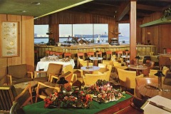A retro-style Sea Wolf Restaurant with wooden decor, round tables, and orange cushioned chairs awaits you. A salad spread adorns the foreground table while the bar in the background hosts lined-up stools. Large windows reveal a view of a ship on Jack London Squares shimmering waters.