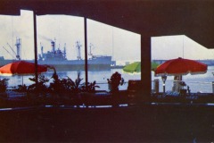A stunning view of the harbor at Jack London Square unfolds through large windows. Inside, red and green umbrellas shade cozy tables. Outside, a ship is docked with cranes silhouetted in the background, while potted plants add a touch of greenery to the scene.