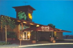 Exterior of Sea Wolf Restaurant at dusk, located in Jack London Square. A neon sign and maroon awning proudly display the restaurants name. The building showcases a modern design with glass doors and surrounding trees, set against a deep blue sky.