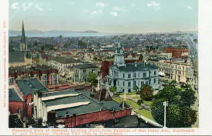 A vintage postcard showing a panoramic view of Oakland, California. The foreground features the historic City Hall with a clock tower, surrounded by smaller buildings and streets. The background displays a sprawling urban landscape under a partly cloudy sky.